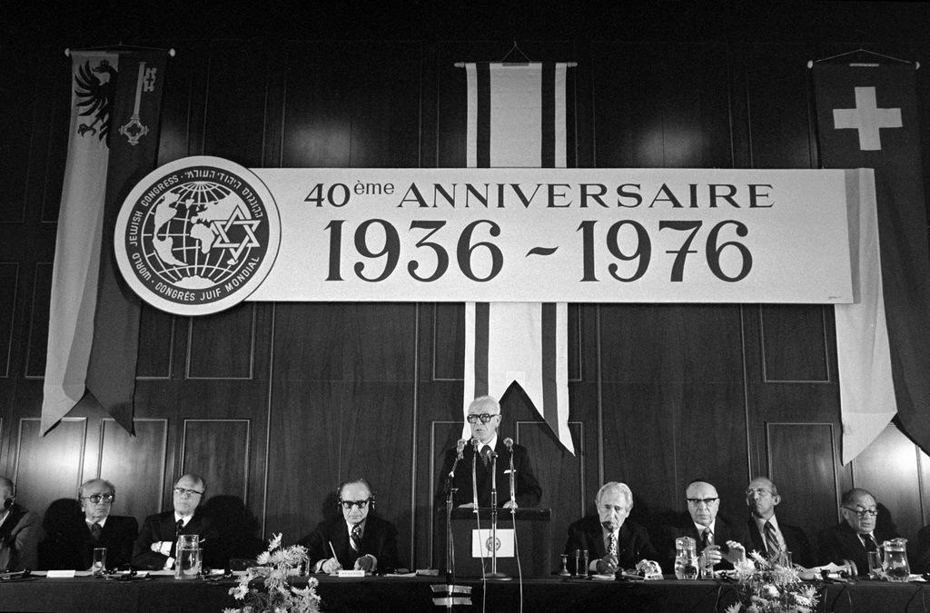Oratori a un tavolo sotto bandiere di Ginevra e Svizzera e la scritta 40° anniversario 1936-1976