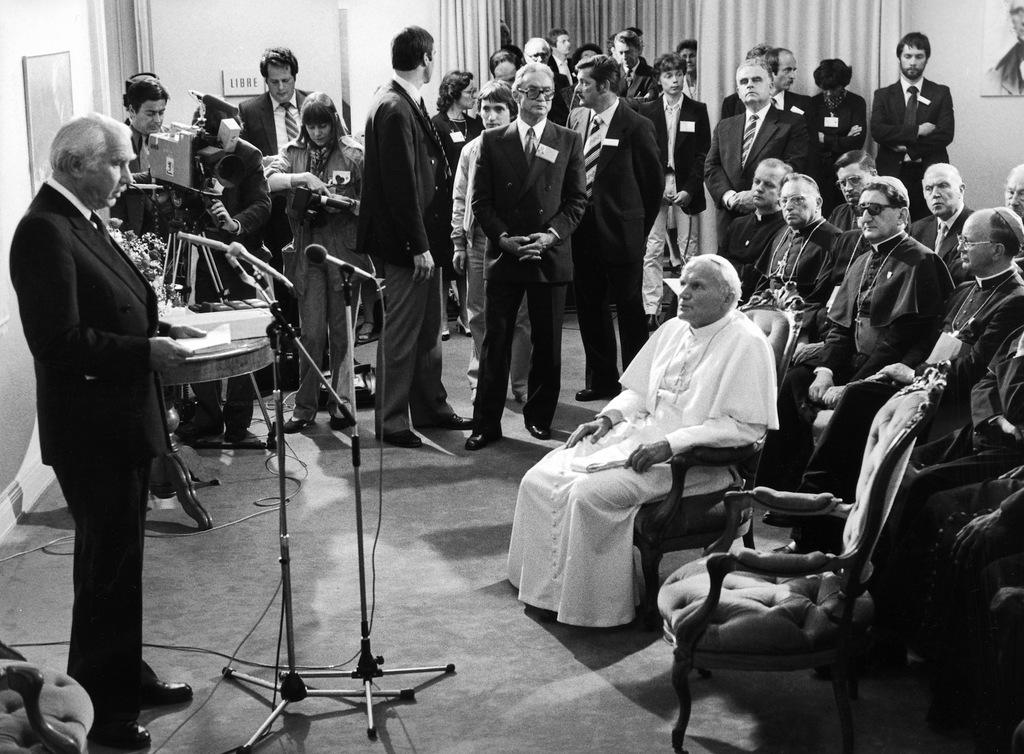Papst hört sitzend einem stehenden Mann bei einer Ansprache über Mikrofon zu