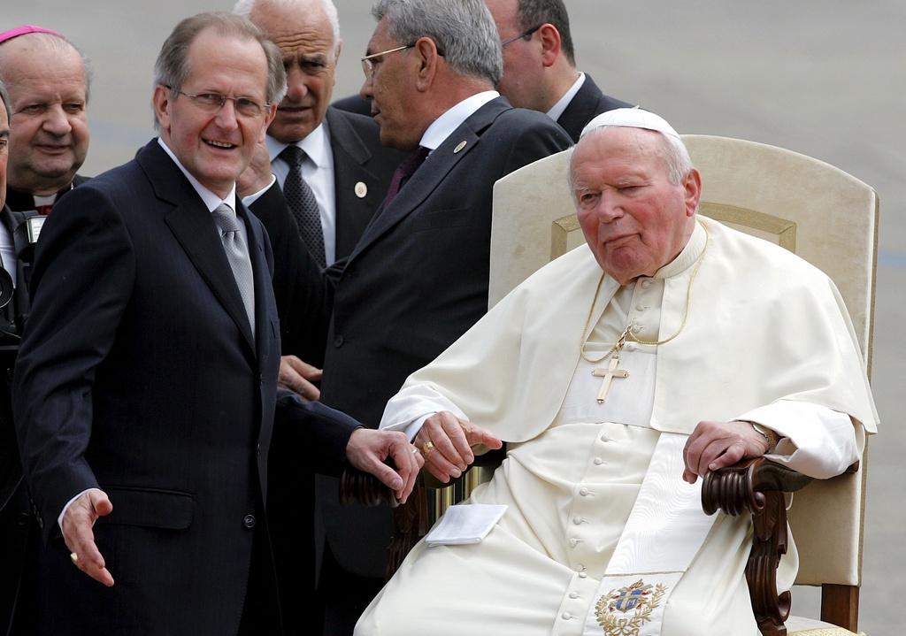 Two men, one to the right in papal regalia of white sitting down.