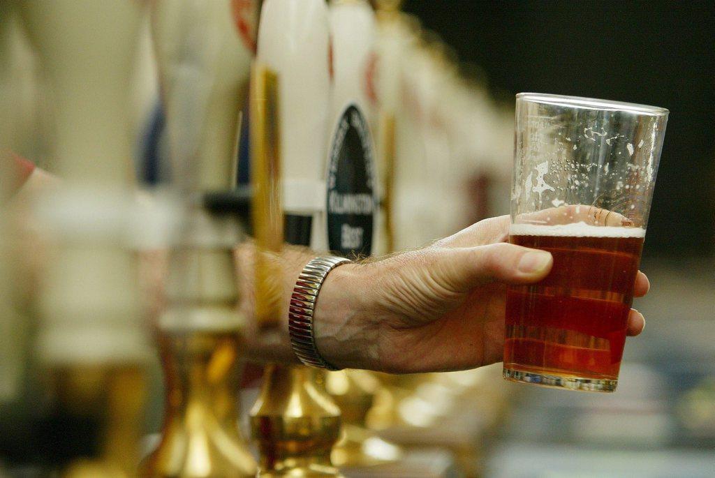 Un bicchiere di birra a metà, mostrato da un uomo del quale si vede solo l avambraccio tra gli stantuffi di un bancone