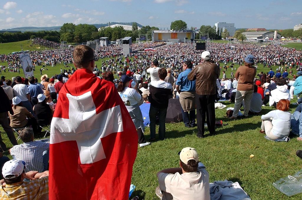Person in the foreground wears a Swiss flag, behind; a field ful of people.