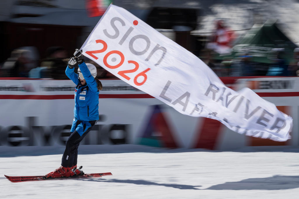 Una niña, en una pista de esquí, lleva una banderola con la inscripción 2026