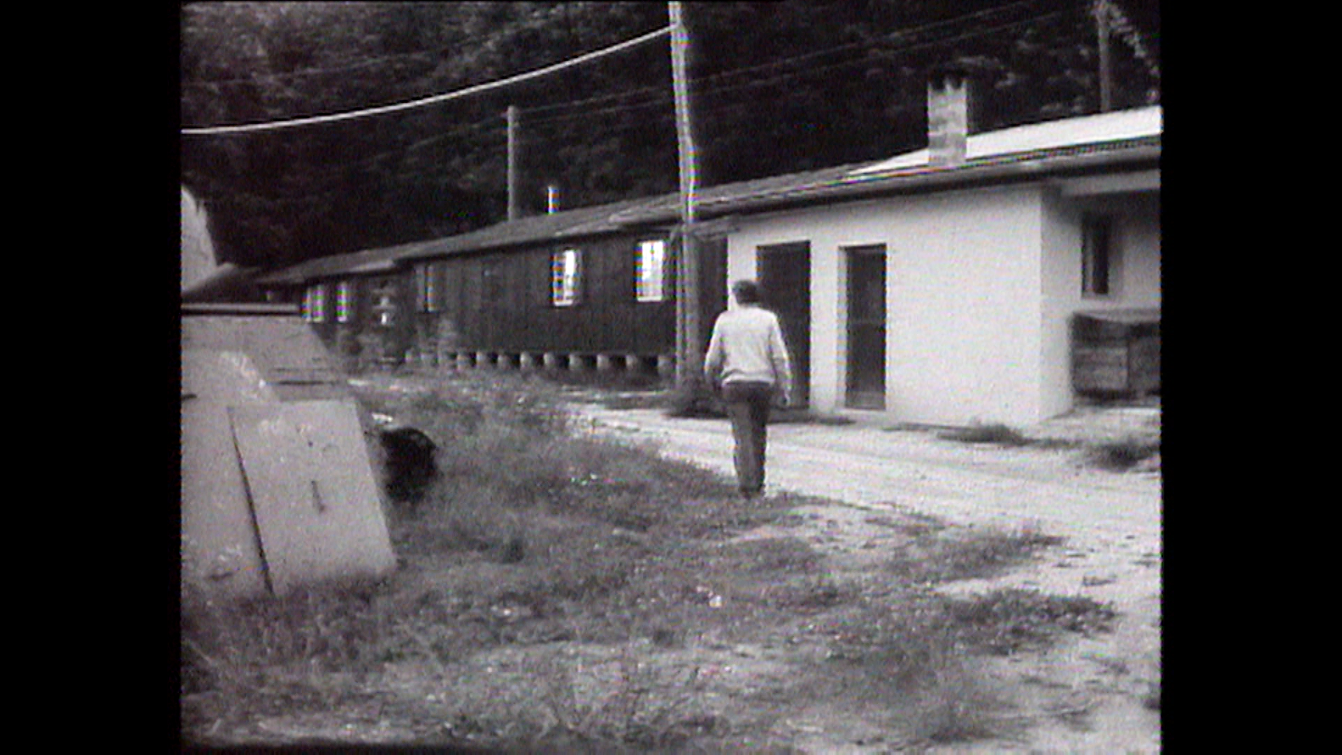 Uomo cammina verso un fabbricato metà di cemento metà in legno; linee elettriche aeree; prato incolto davanti