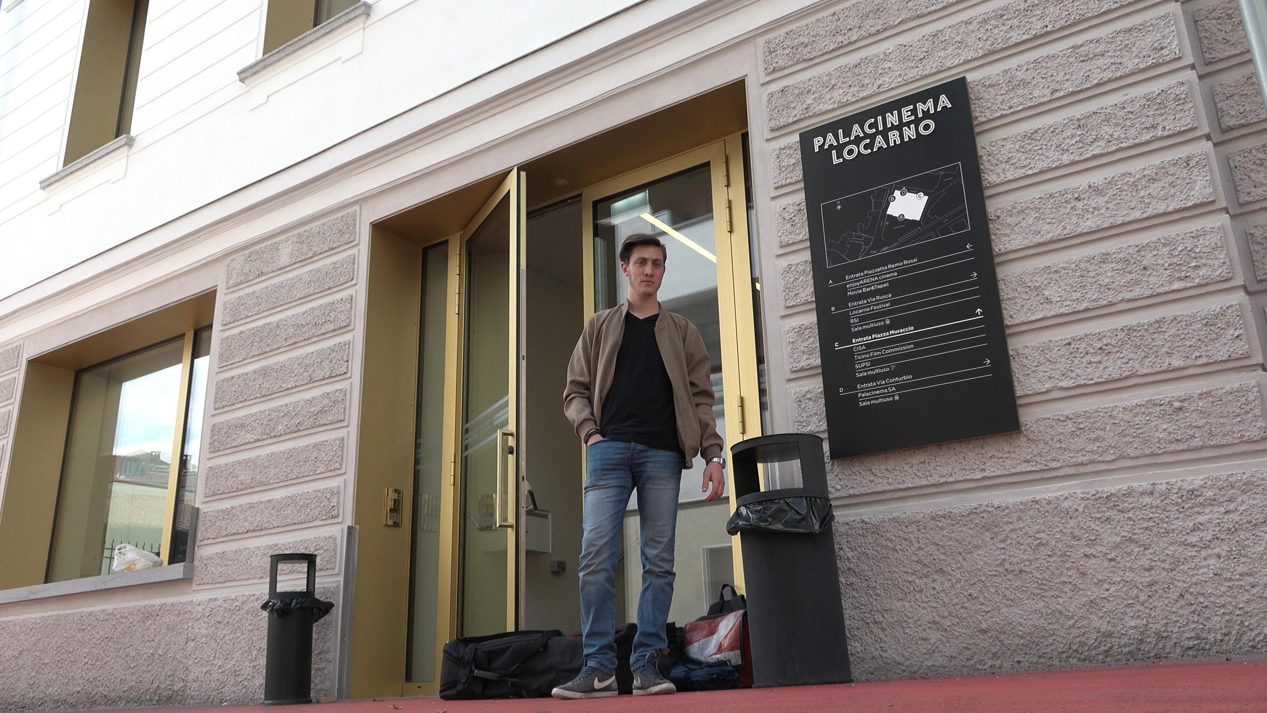 Ein junger Manns steht vor einer halboffenen Türe, zu seinen Füssen liegen Taschen.