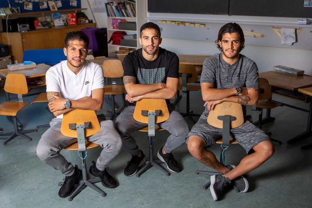 Drei junge Männer sitzen in einer Schulstube auf Stühlen und schauen in die Kamera.