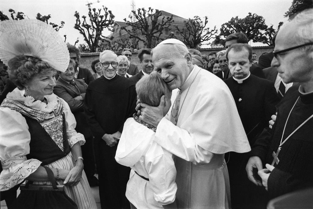 A man in papal regalia embraces a young person while meeting people.