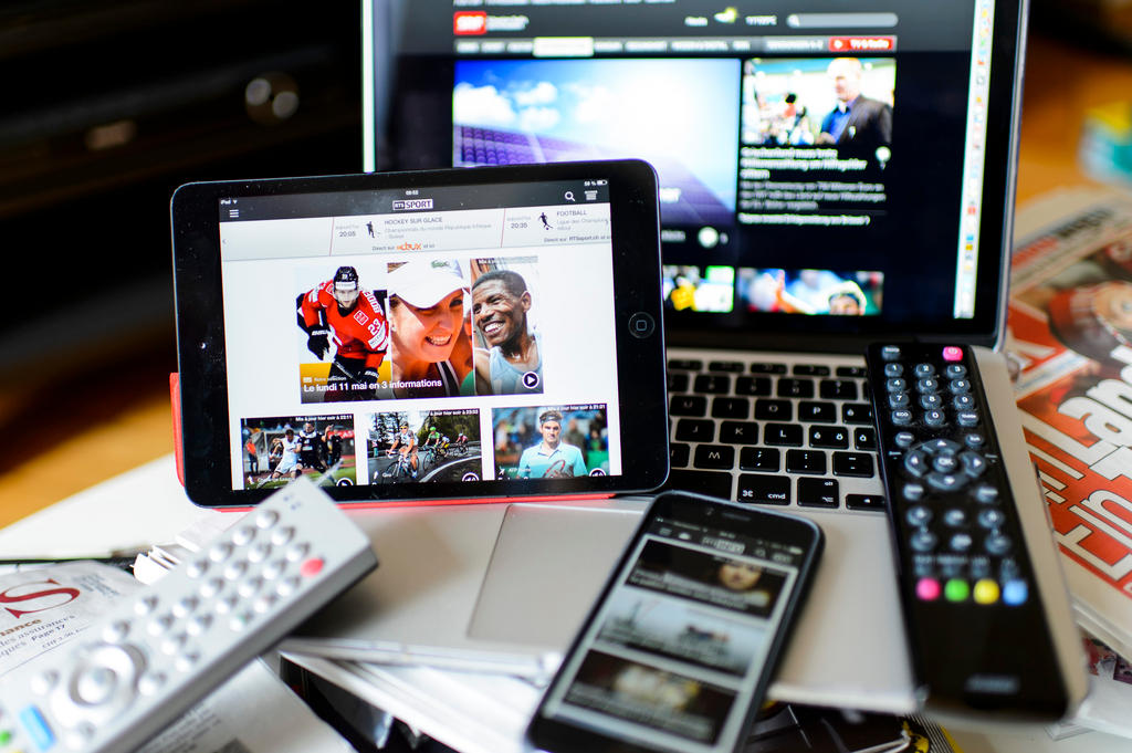 Laptop screen, tablet, mobile phone, remote controls and newspaper on a table