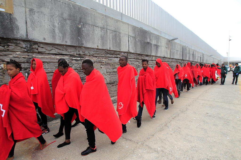Fila di profughi con coperte rosse cammina lungo un muro