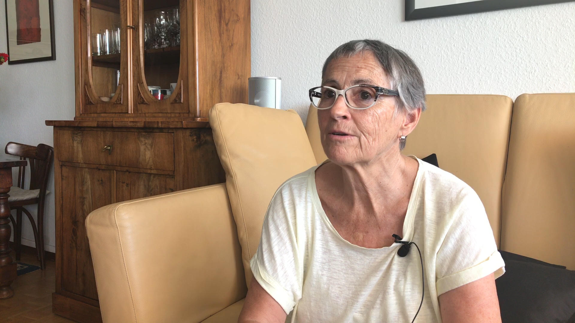Eine ältere Frau sitzt auf einem hellbraunen Sofa und schaut in die Kamera.