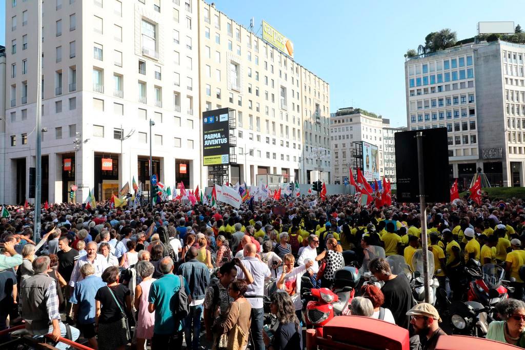 Vista di piazza San Babila a Milano colma di persone; bandiere rosse in lontananza