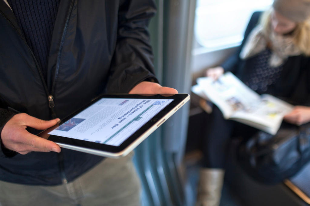 A man rides the tram while reading a newspaper on his iPad.