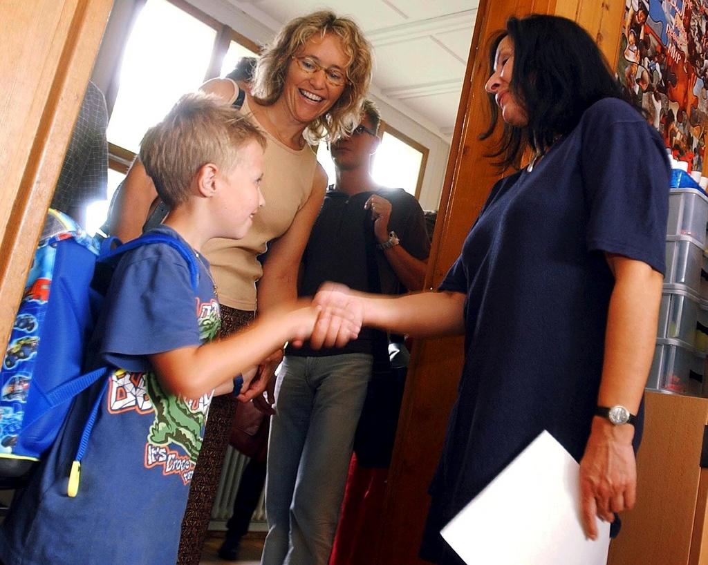 un bambino stringe la mano a una maestra, mentre la sua mamma lo osserva sorridendo.