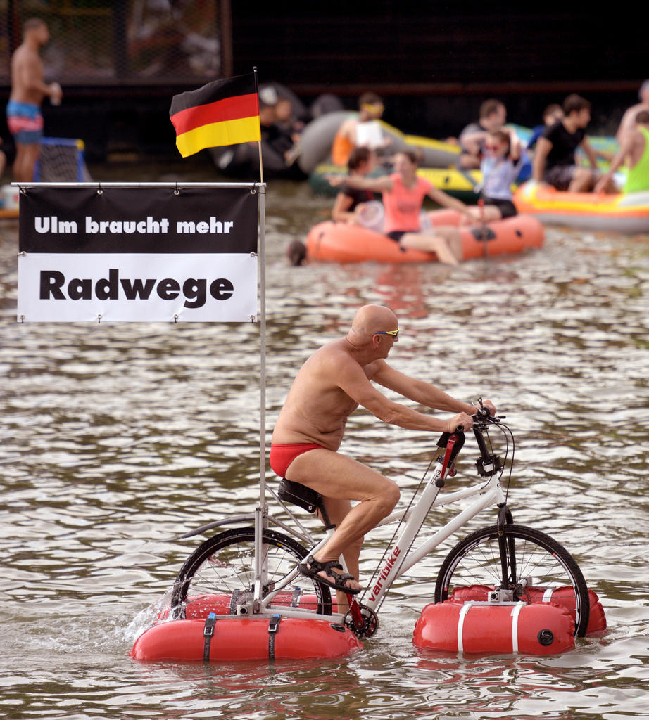 Mann fährt mit Fahrrad, an dem Schwimmer angebracht sind, auf Fluss mit Transparent: Ulm braucht mehr Radwege