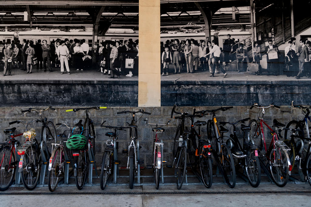 Geparkte Fahrräder vor einem Fotografie schwarzweiss von wartenden Menschen an einer Bahnstation in Japan.