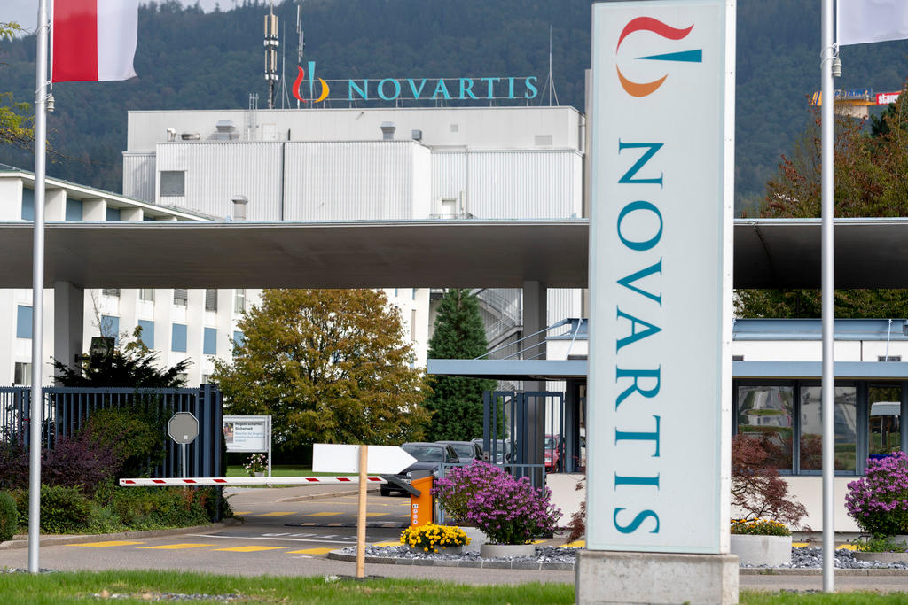 Totem di Novartis in primo piano, dietro di esso un accesso veicolare a uno stabilimento industriale con l insegna Novartis