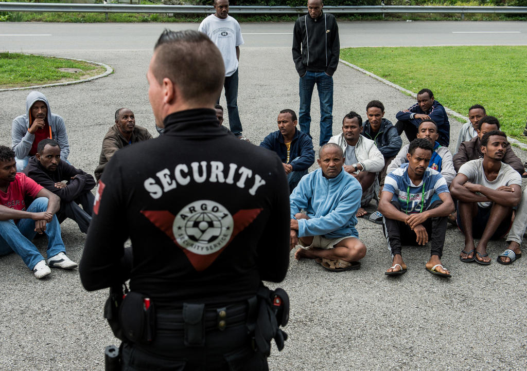 A group of seated Eritrean asylum seekers are watched over by a security guard