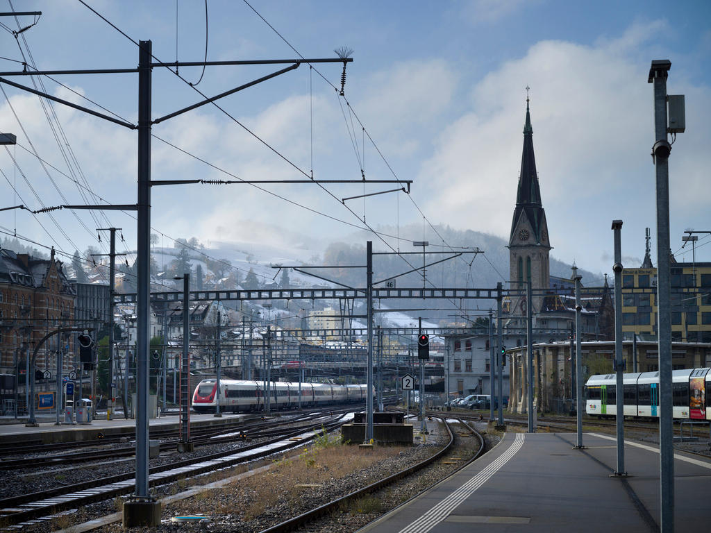 Immagine di stazione ferroviaria; in primo piano banchine e binari; due convogli in arrivo; cittadina sul fondo