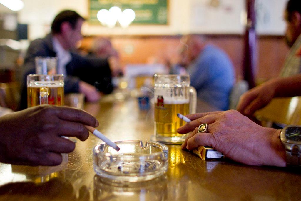 Primo piano di due boccali di birra, un portacenere e due mani che reggono una sigaretta ciascuno; sul fondo ambiente bar sfocat