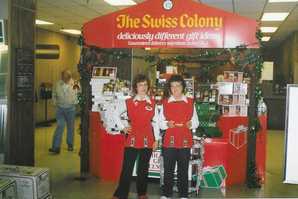 Two saleswomen stand in front of a Swiss Colony stand, in red aprons