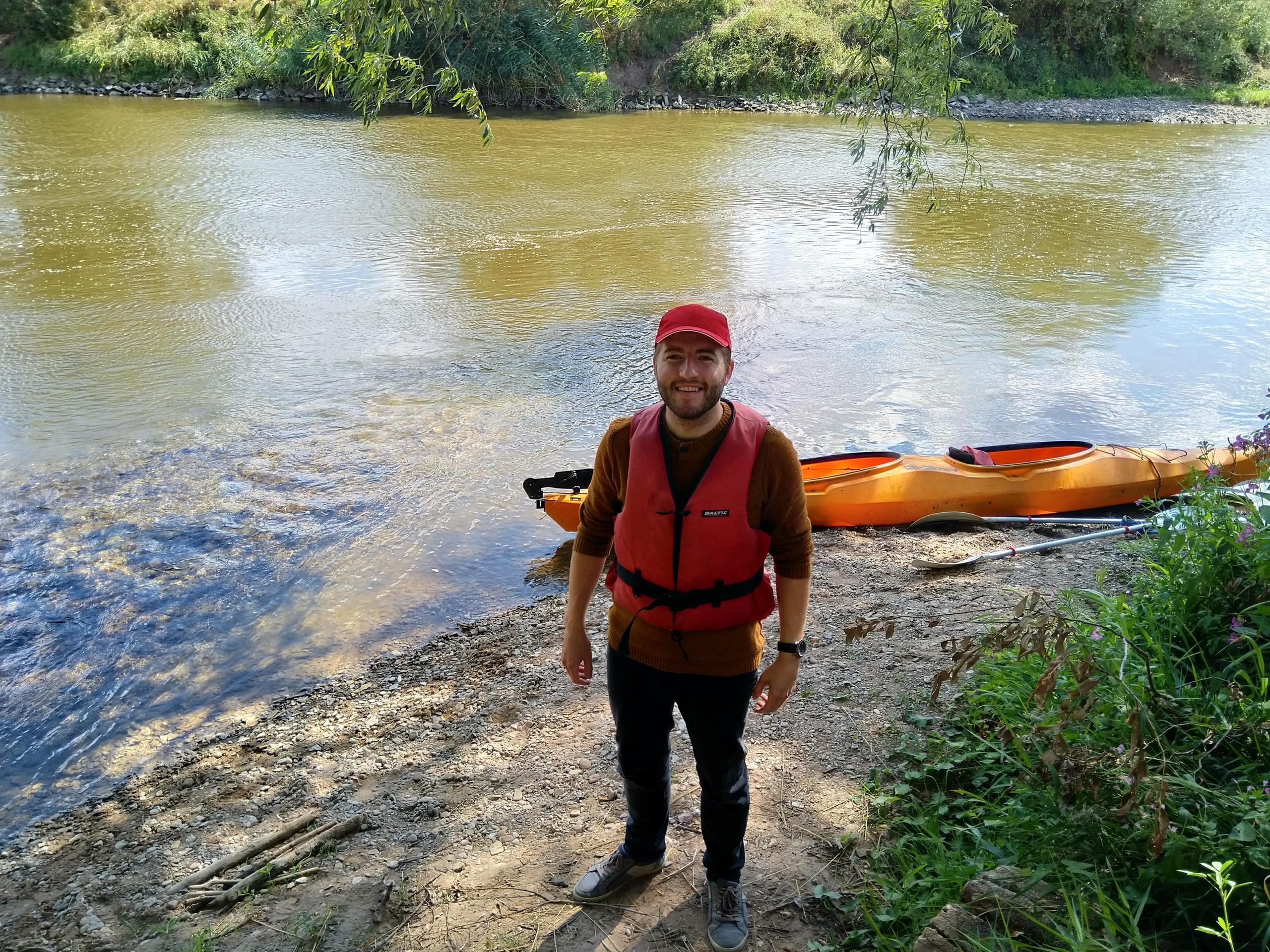 Mann in Schwimmweste am Ufer eines Flusses, dahinter ein Paddelboot