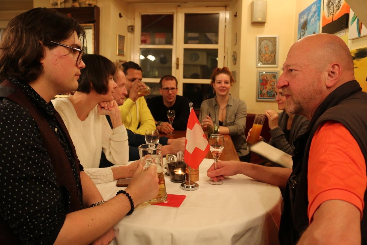 Menschen sitzen in einem Lokal um einen Tisch und diskutieren, in der Mitte steht eine Schweizer Fahne.