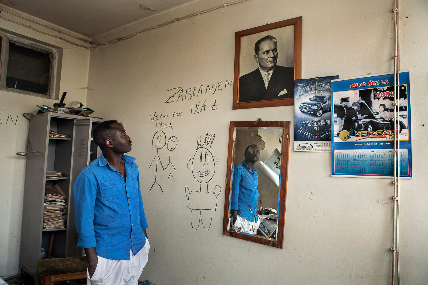 A man stands in a roo with his hands in his pockets looking towards a picture on the wall.