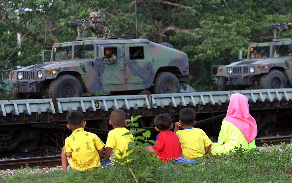 Kinder sitzen an einem Strassenrand im Gras und schauen zu, wie Panzerfahrzeuge vorbeifahren.