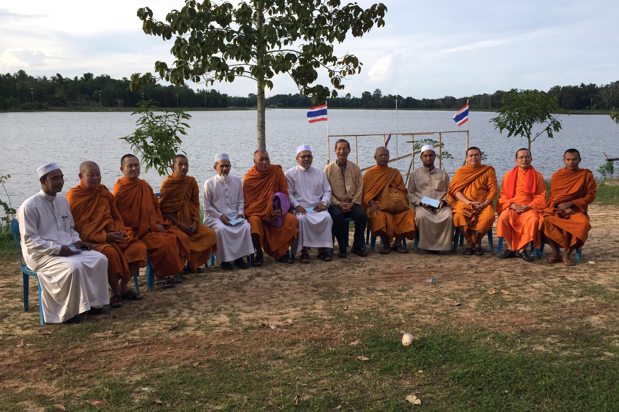 Un gruppo di uomini, buddhisti e musulmani, è seduto a semicerchio su un prato con il mare alle spalle.