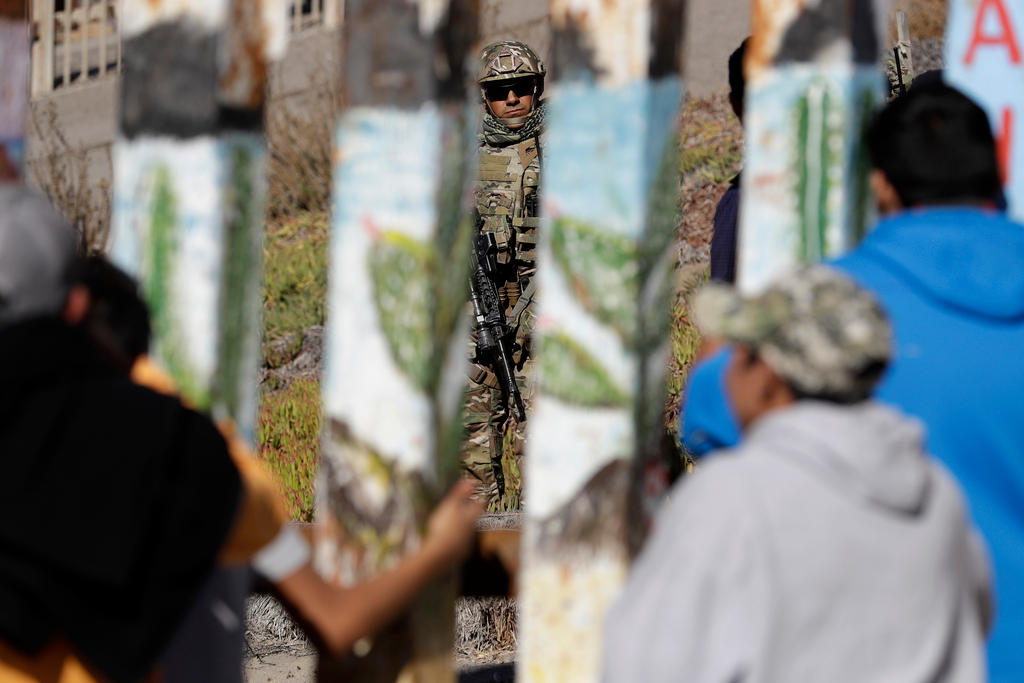 Migranti di spalle e fitte colonne di cemento, in primo piano ma sfocati; si intravede (a fuoco) un militare armato oltre il mur