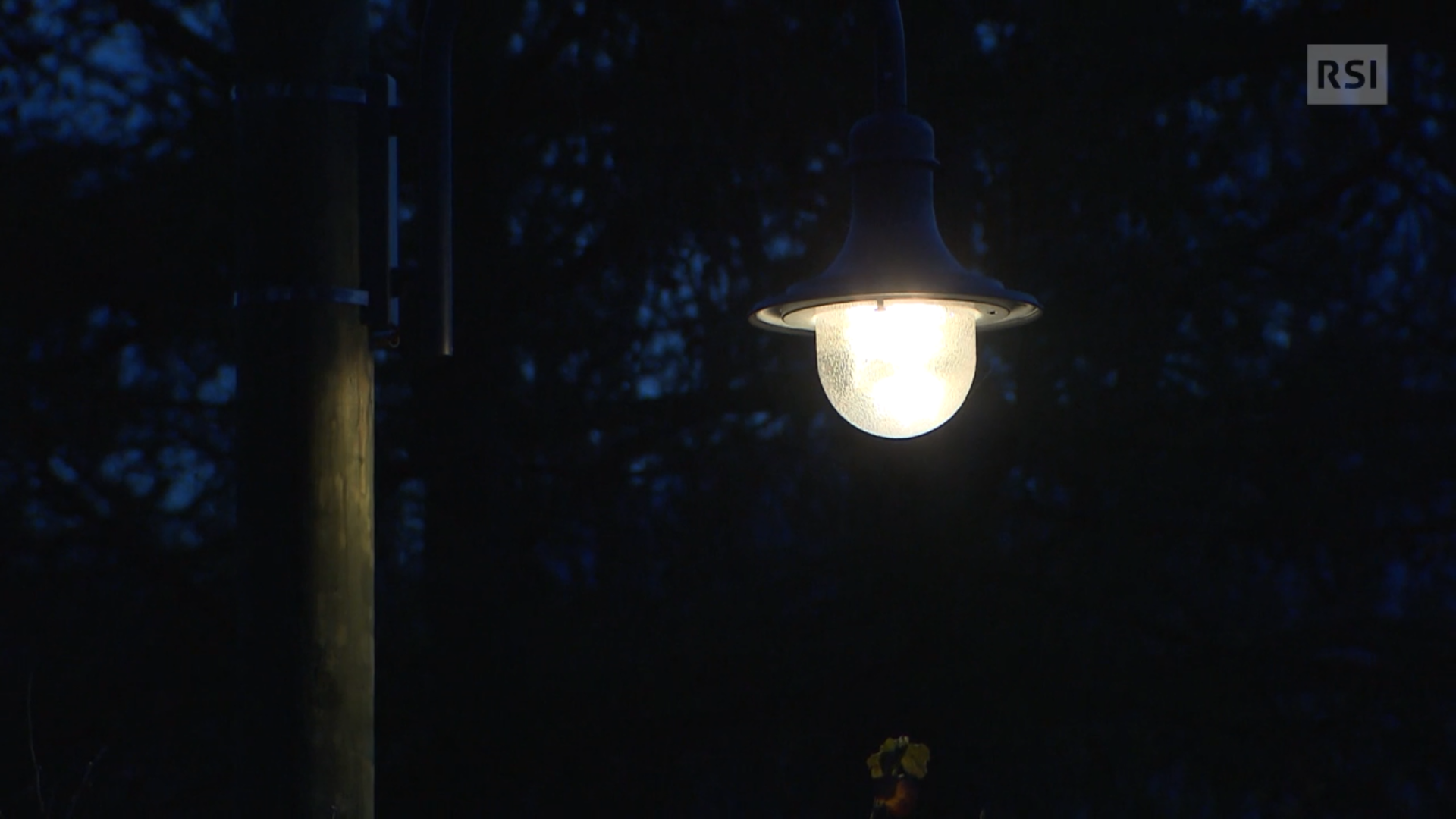 Primissimo piano di un lampione a lanterna; sulla sx palo flebilmente illuminato