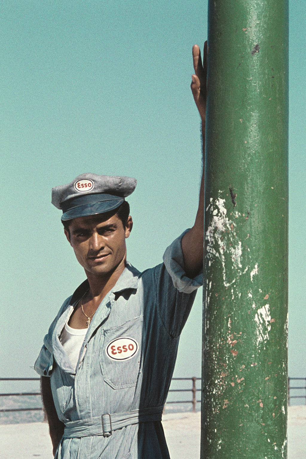 Homem vestindo um uniforme de posto de gasolina Esso, apoiado a um poste