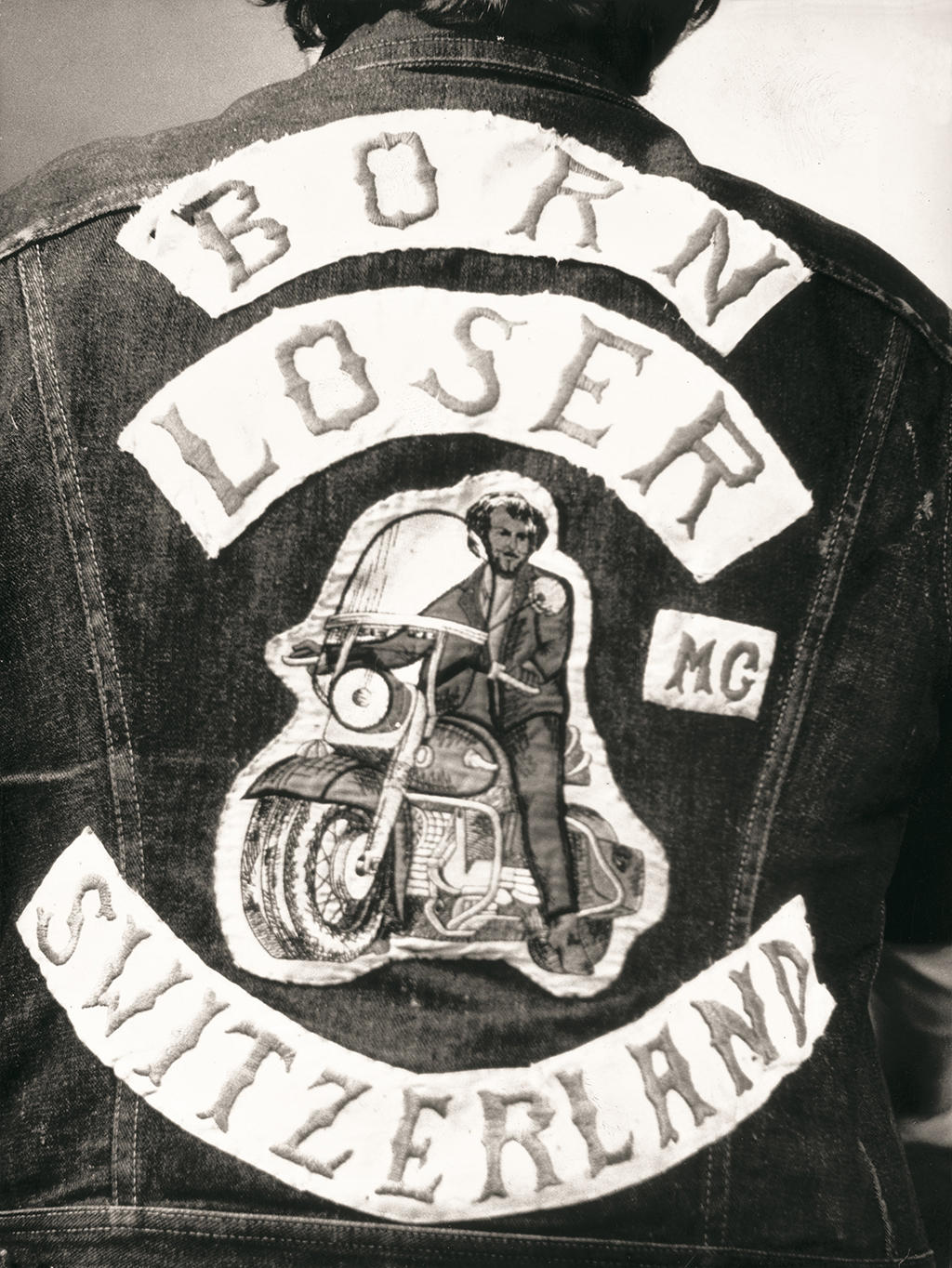 Sewn on patches on the back of a denim jacket which reads Born Loser Mc Switzerland.