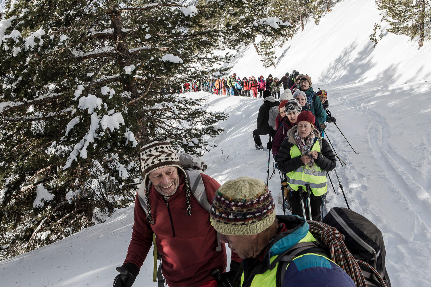 A line of people walk up a snowy alpine terrain towards the camera, dressed in winter clothes.