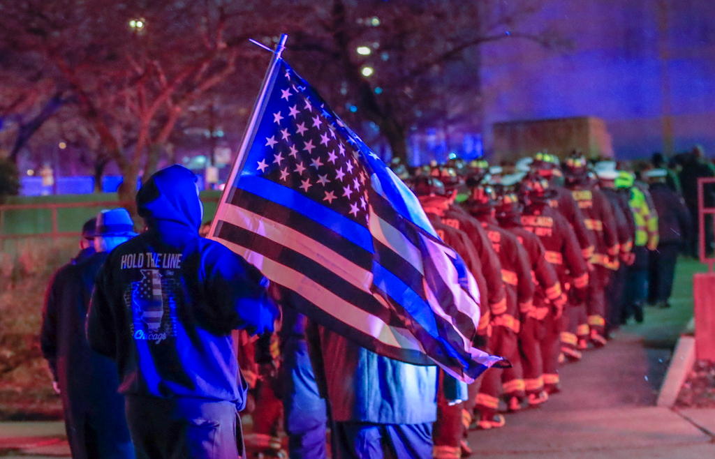 Pompieri in fila davanti all ospedale e alla fine della fila un uomo porta la bandiera americana