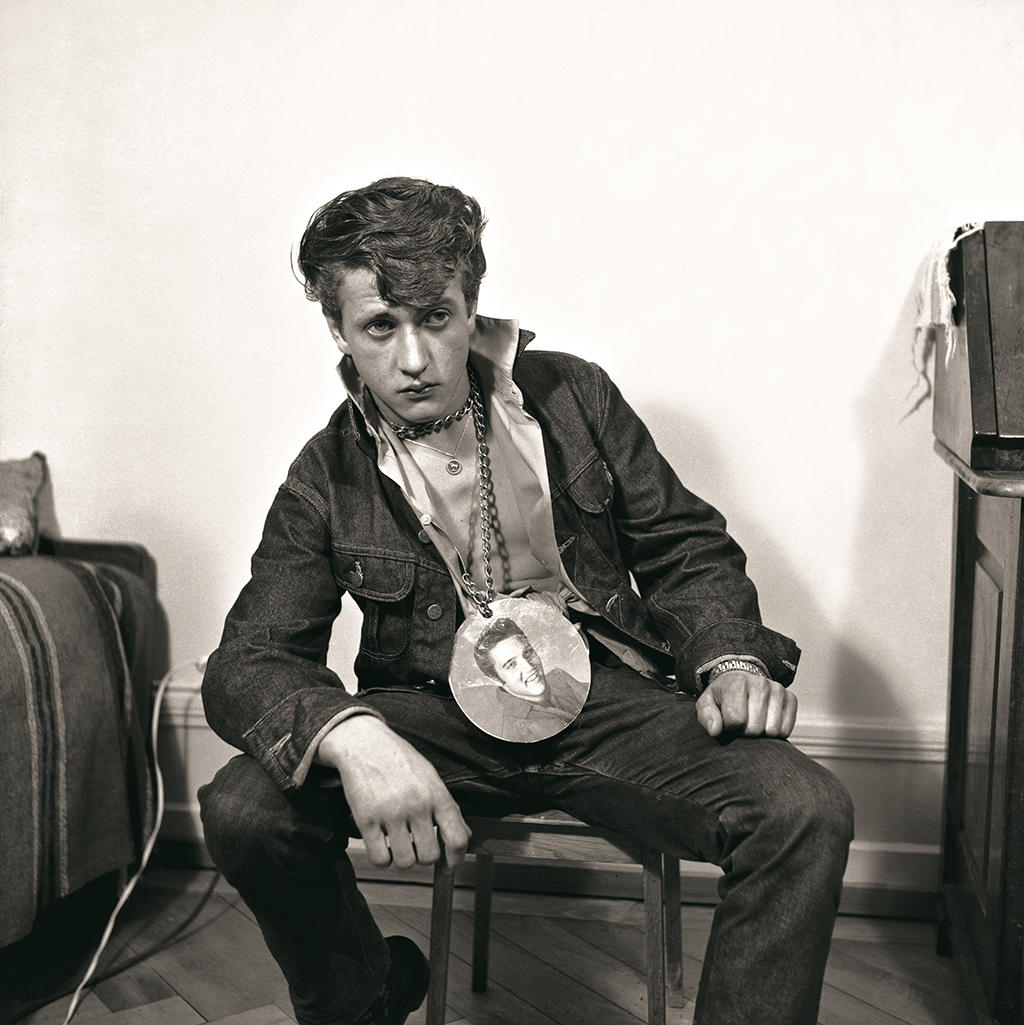 A man sits leaning, with an image of Elvis on a chain around his neck.