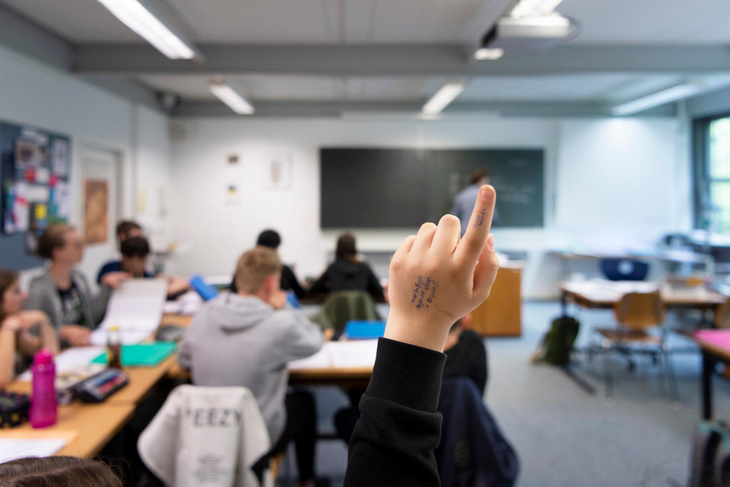 Immagine sfocata di una classe di scuola, sul fondo insegnante e lavagna; in primo piano, a fuoco, mano di un allievo alzata