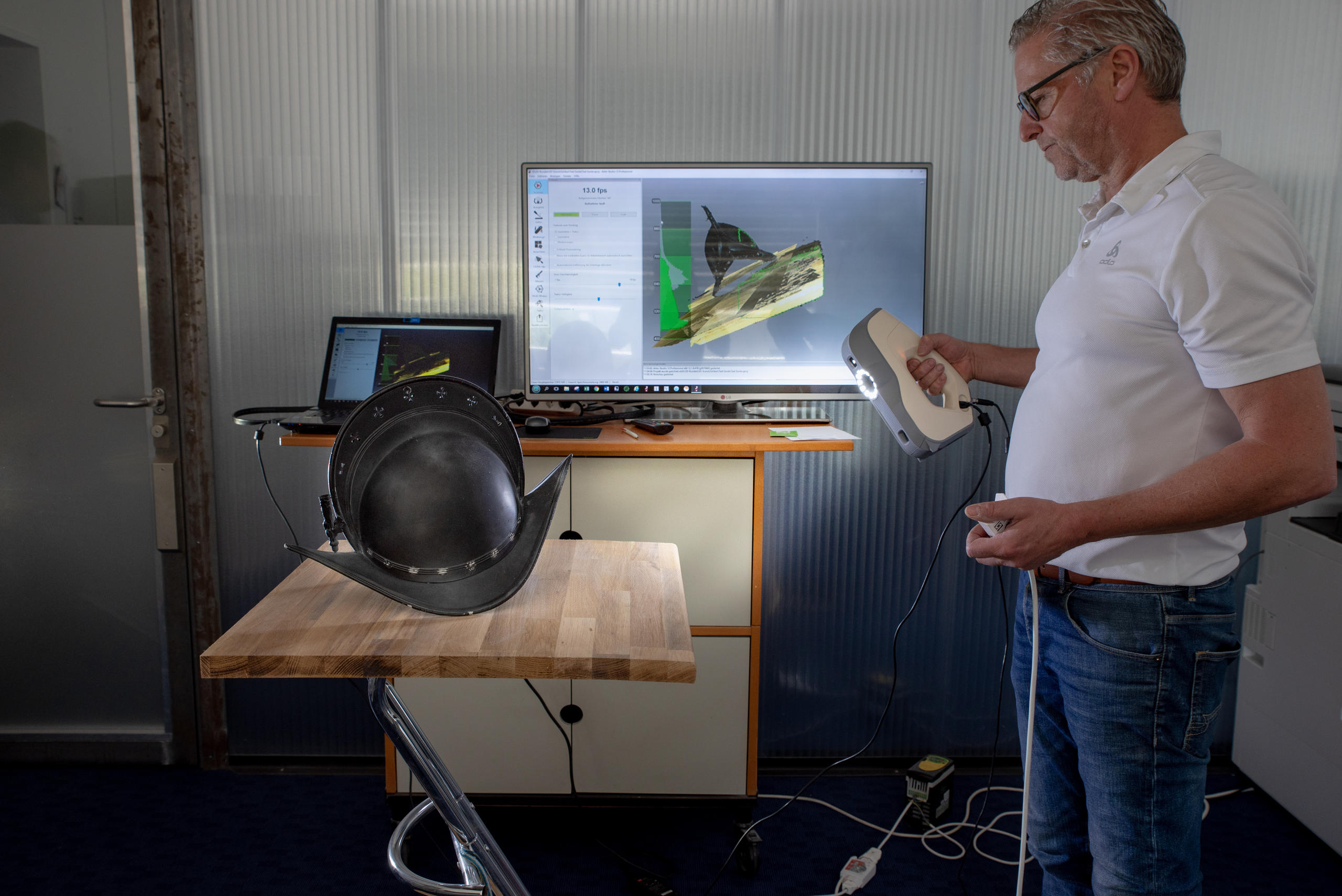 Homem escaneia um capacete sobre uma mesa e um monitor de vídeo no fundo.