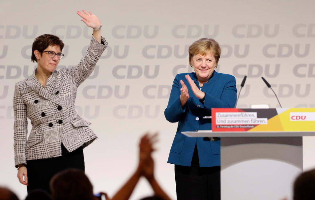 Palco con scritte CDU sul fondo; sulla sx Kramp Karrenbauer alza la mano in segno di saluto; sulla dx Merkel applaude e sorride
