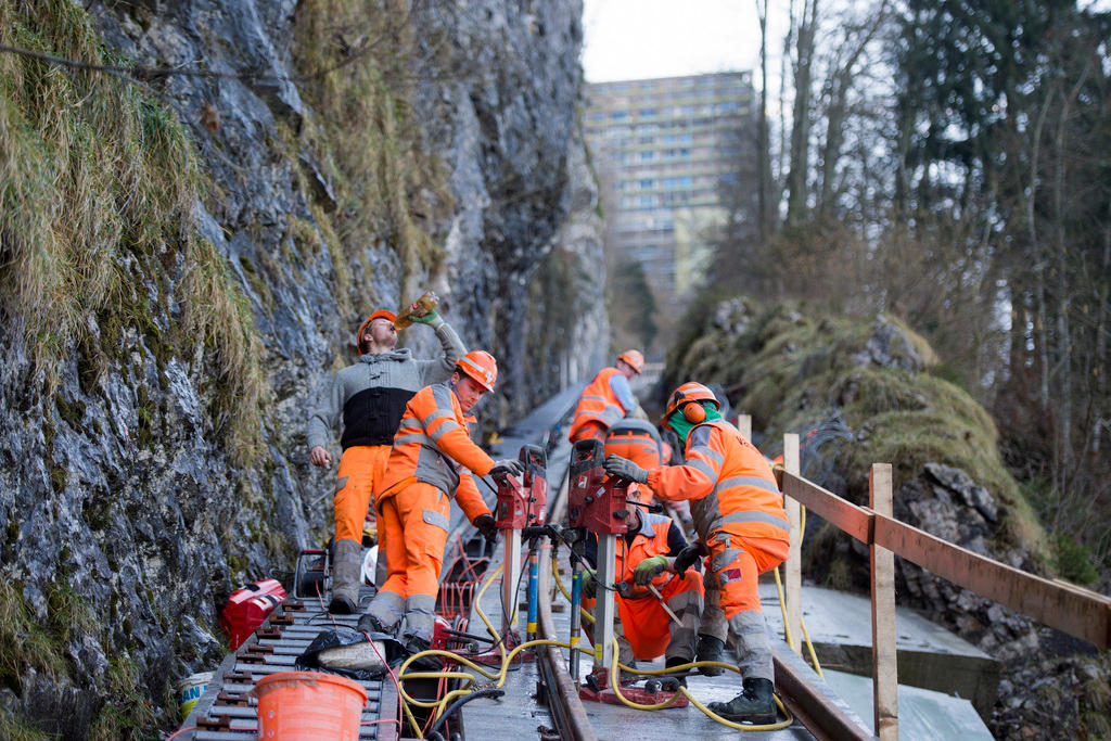 Männer in orangen Anzügen und Schutzhelmen auf einer Baustelle unter einer Felswand