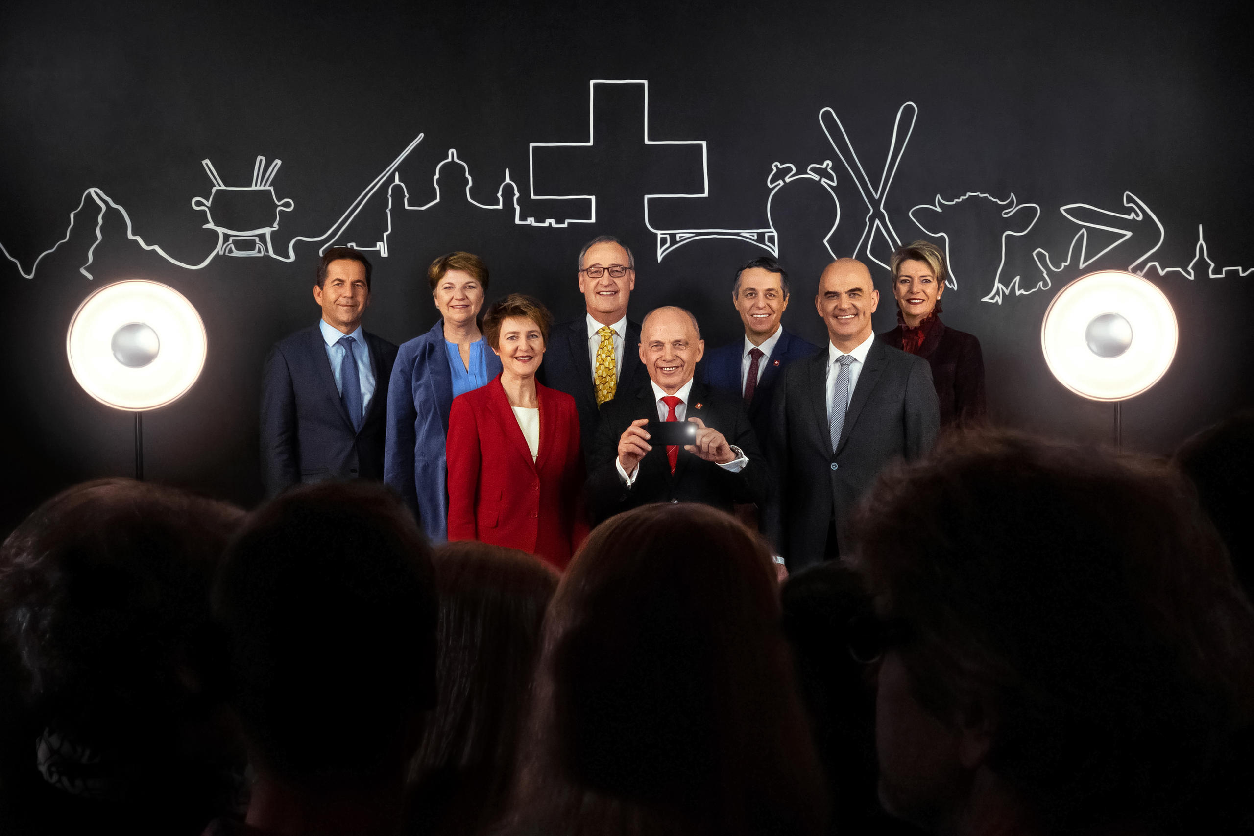 Foto di gruppo del governo (Maurer, al centro, regge uno smartphone); dietro, simboli stilizzati; davanti, persone di spalle