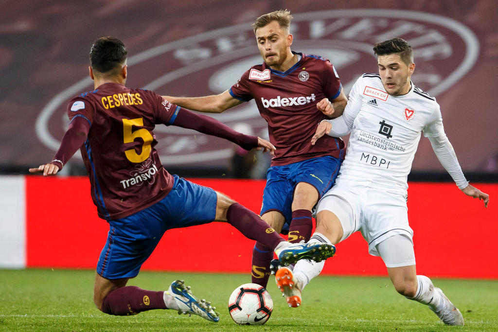 Drei Fussballer, zwei im roten, einer im weissen Dress, kämpfen um einen Ball