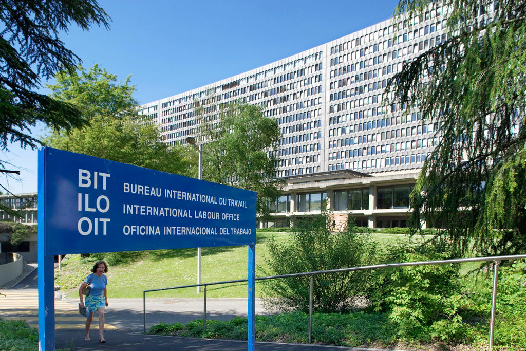 Ein blaues Schild steht vor einem grossen, langen Gebäude, das viele Fenster hat.