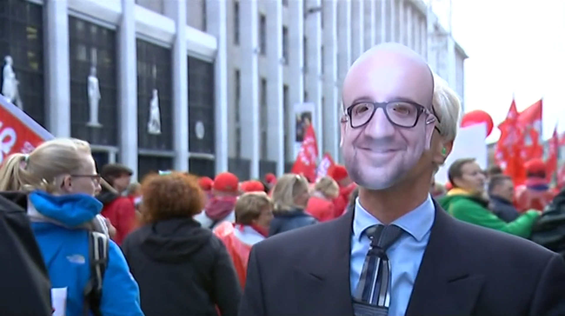 Primo piano di un uomo che indossa una maschera col volto di un altra persona, all interno di un corteo con bandiere rosse