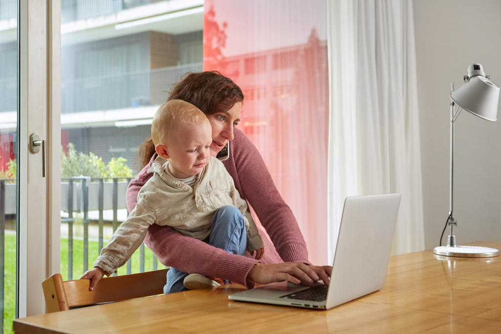 Una donna a casa davanti al computer mentre telefona e ha in braccio un bimbo piccolo