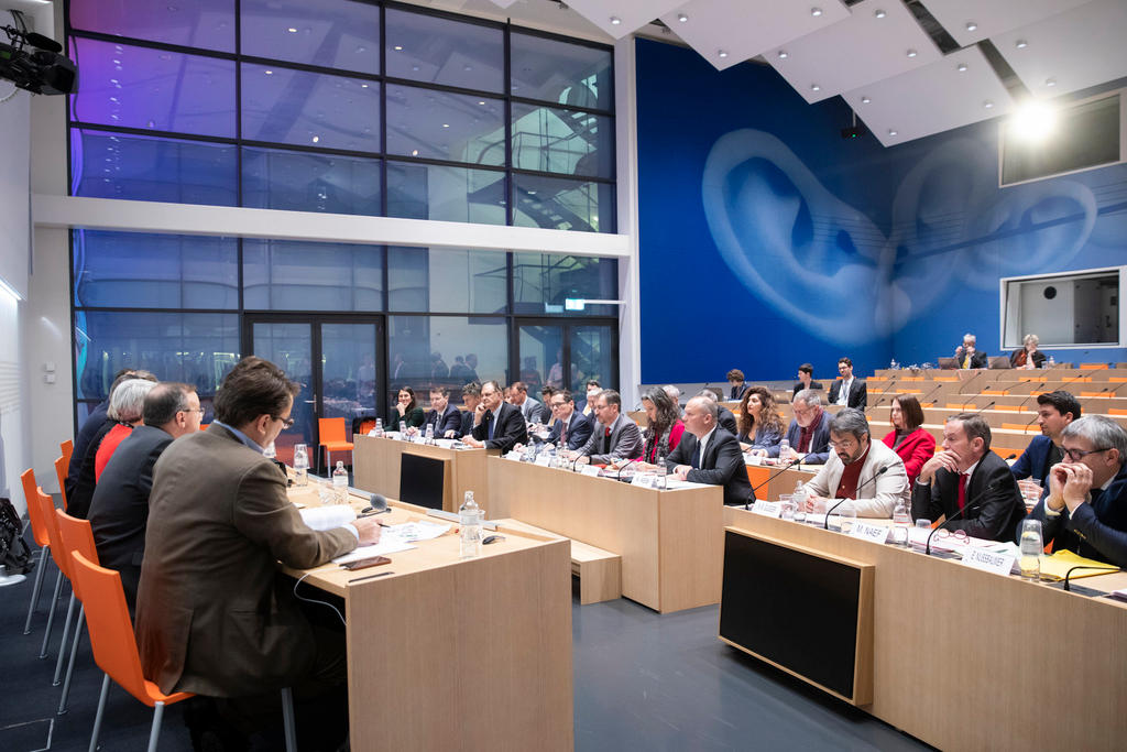 Vista generale di una sala stampa con platea occupata per metà, grandi vetrate e un orecchio dipinto alle pareti