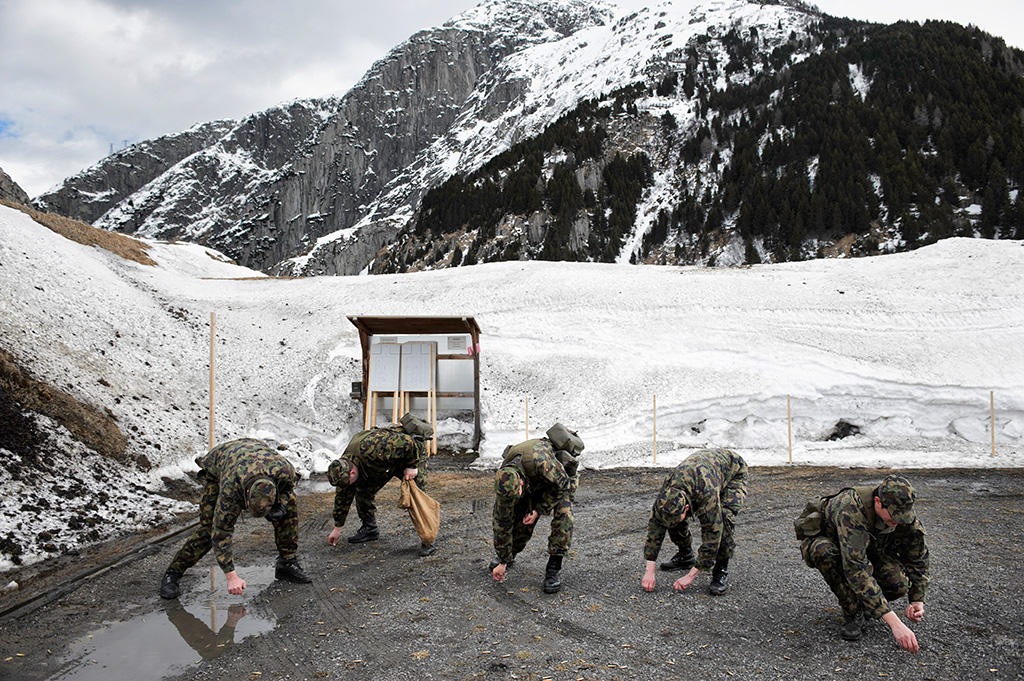 Uomini con uniforme mimetica raccolgono qualcosa da terra in un paesaggio sterrato in parte innevato