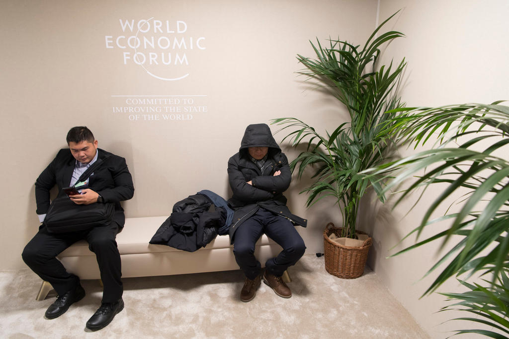 Two men sit on a bench inside a room with plants, one of the men wears a jacket with the hood over his head.