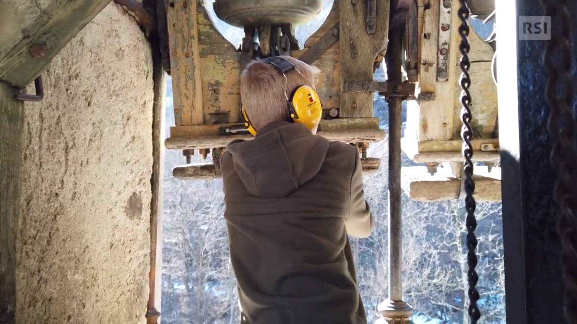 Ragazzino di schiena in una cella campanaria; indossa cuffie di protezione e sta per mettere in moto una campana