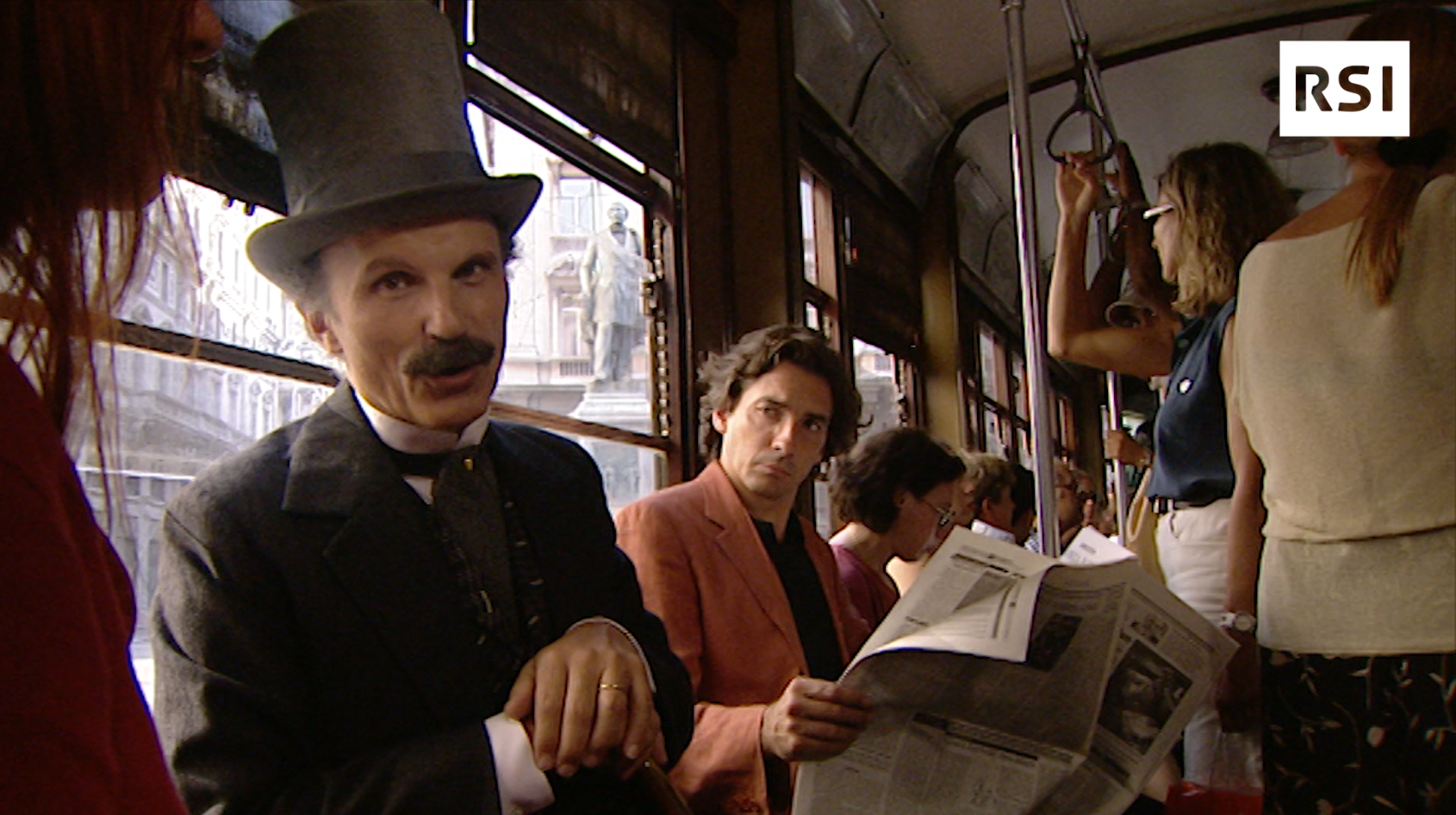 Un uomo vestito stile 800, seduto in un tram, guarda e parla verso la camera; un vicino lo osserv