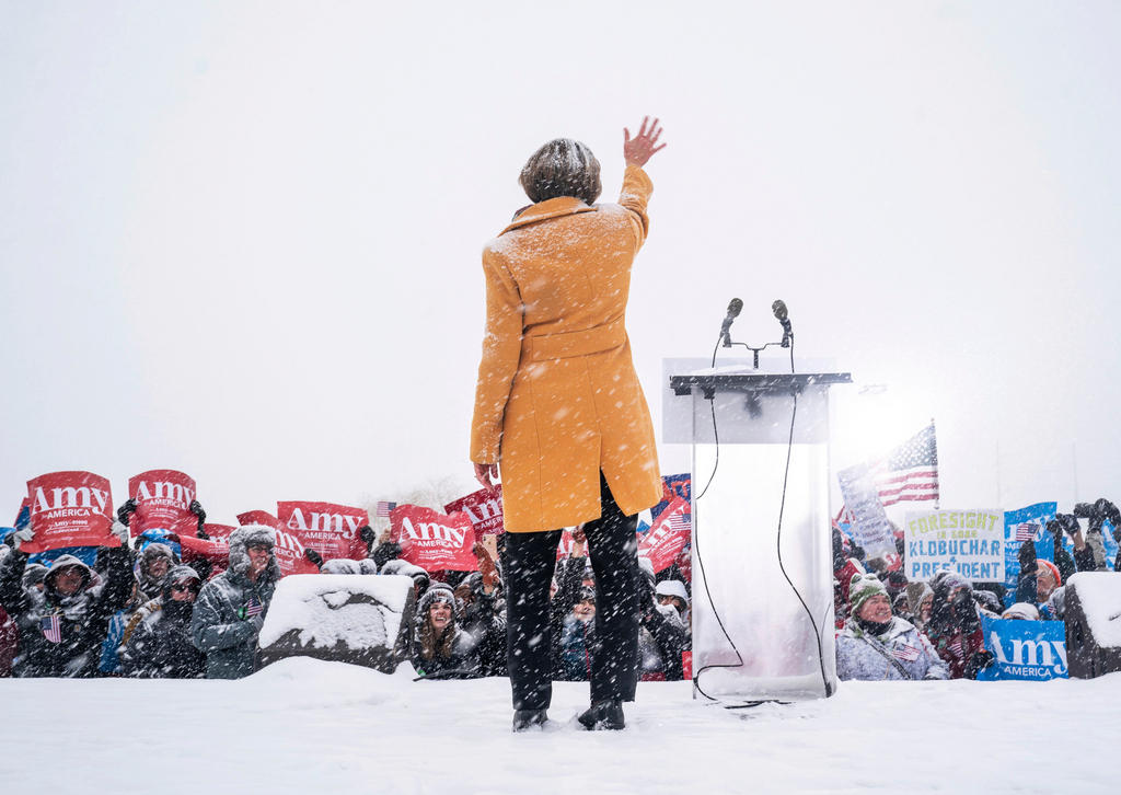 Una donna, di schiena, accanto a un pulpito con microfoni saluta folla sotto la neve che regge cartelli con scritto Amy